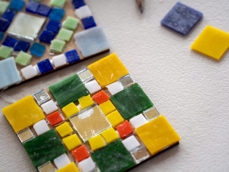 Image of Two colourful mosaic coasters made of square glass tiles in yellow, green, blue, white, and orange are arranged on a light surface, with loose tiles nearby.