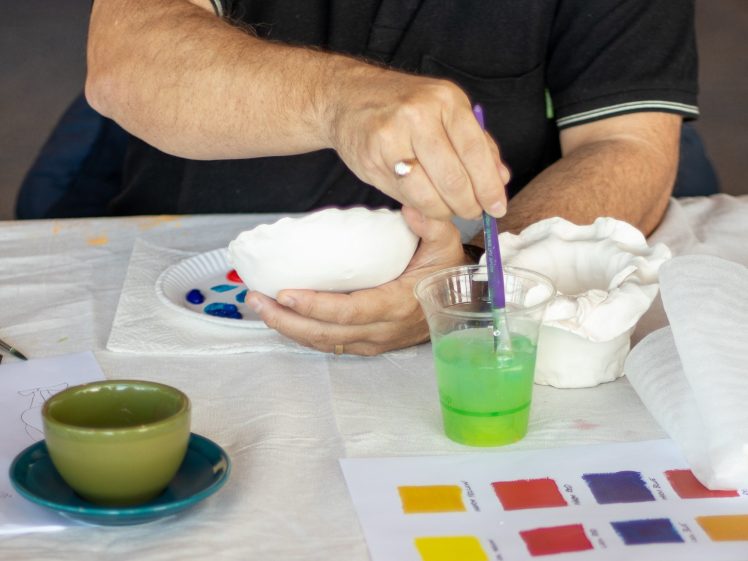 Image of A person holds a white ceramic bowl while dipping a paintbrush into a cup of green water. On the table are paint, a ceramic piece, a green teacup, and a colour chart.