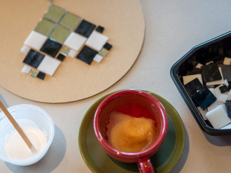 Image of A red coffee cup on a green saucer sits next to a small bowl of white glue, a wooden stick, mosaic tiles on a round board, and a tray of loose black, white, and green tiles on a table.