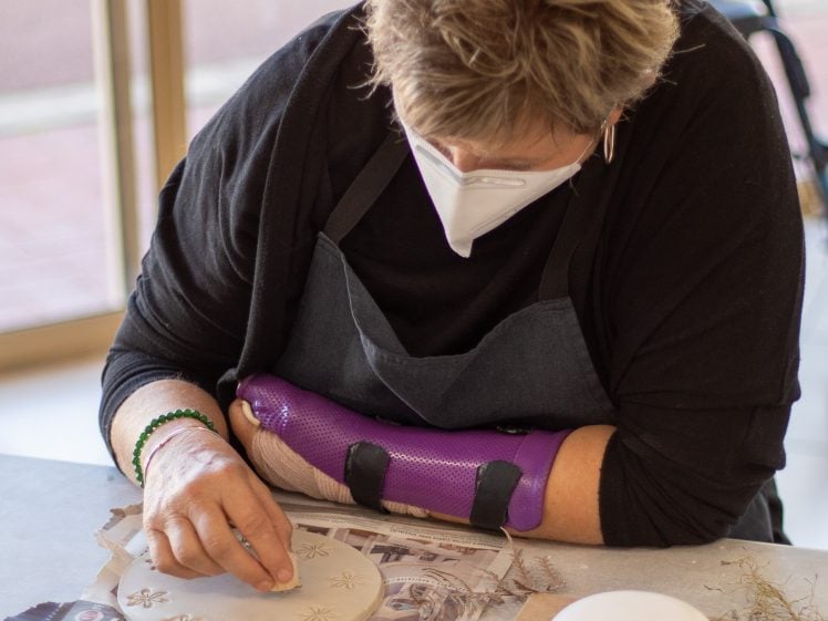 Image of A person wearing a mask and purple arm support decorates a circular clay piece with star designs at a table covered with tools and materials.