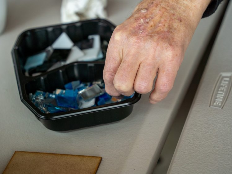 Image of A hand reaches towards a tray filled with blue and clear glass pieces on a light grey table, with a small piece of wood and a “LIFETIME” logo visible nearby.