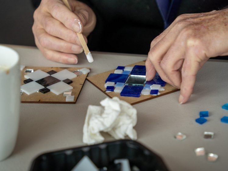 Image of Close-up of hands creating mosaic art with blue, white, and silver tiles on brown boards. Tools, loose tiles, a white cup, and a tissue are on the table.