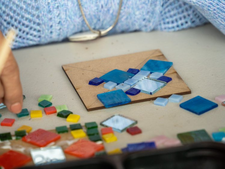 Image of A person arranges colourful glass tiles on a square wooden board, likely creating a mosaic. The table is scattered with various small, vibrant tiles, and the person wears a blue knitted jumper.