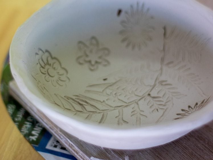 Image of A handmade ceramic bowl with floral and fern leaf patterns etched inside, resting on a wooden surface with some paper and tools nearby.