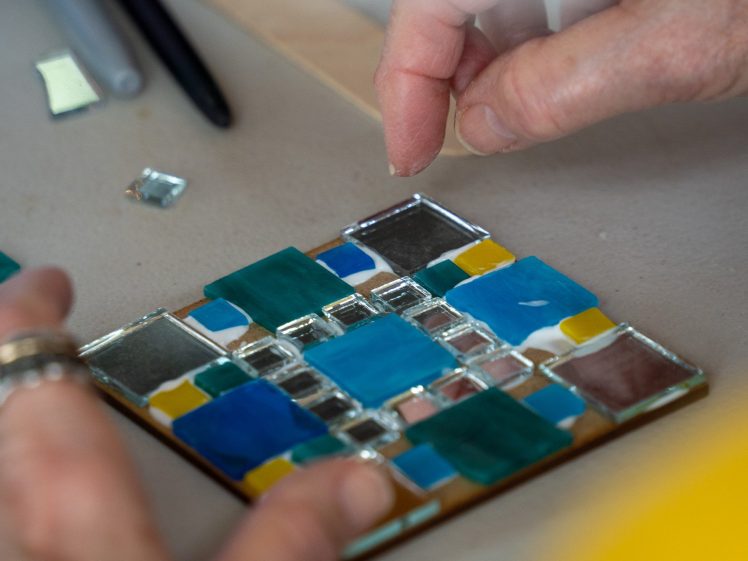 Image of Close-up of hands arranging colourful glass tiles on a square base, creating a mosaic art piece. The tiles are blue, yellow, green, and clear, and the work is being done on a light grey surface.