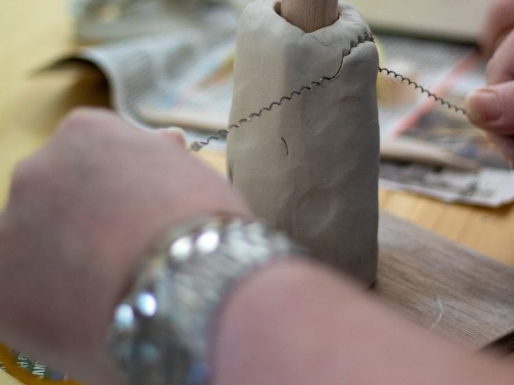 Image of A person uses a wire clay cutter to slice through a block of clay that is held upright on a wooden board. Newspapers are scattered in the background.