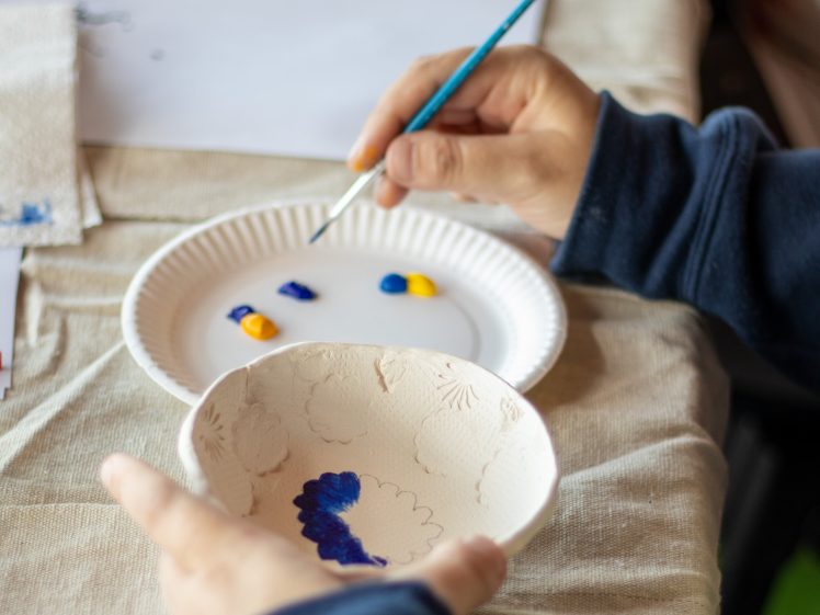 Image of A person holds a ceramic bowl while painting it blue with a small brush. The hand reaches for acrylic paint on a white paper plate palette, resting on a beige tablecloth.