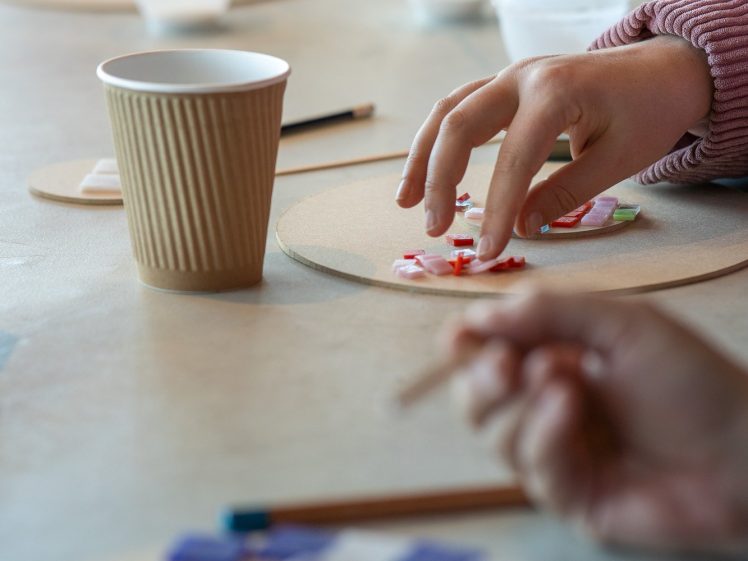 Image of Close-up of hands placing colourful mosaic tiles onto a circular board on a table, with a brown paper cup, pencils, and other craft supplies in the background.