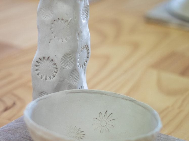 Image of A hand-built clay vase with textured patterns stands behind a round clay bowl with similar designs, both unfired and placed on a wooden board on a light wooden table.