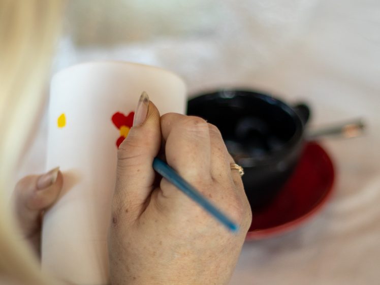 Image of A person holds a white ceramic mug and paints small red and yellow shapes on it with a blue paintbrush. A black cup on a red saucer and art supplies are visible on the table in the background.