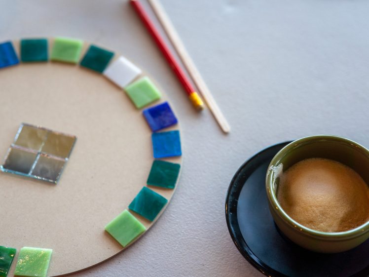 Image of A close-up of a mosaic project with colourful tiles arranged on a circular board, a pair of chopsticks and a red pencil nearby, and a cup of coffee with crema on a black saucer to the right.