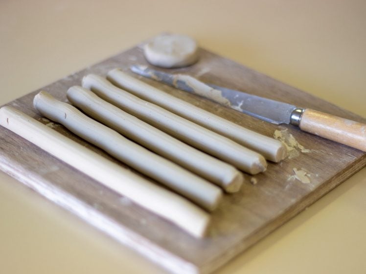Image of Several rolled clay strips and a round clay piece are placed on a wooden board next to a metal knife with a wooden handle, all set on a light-coloured surface.