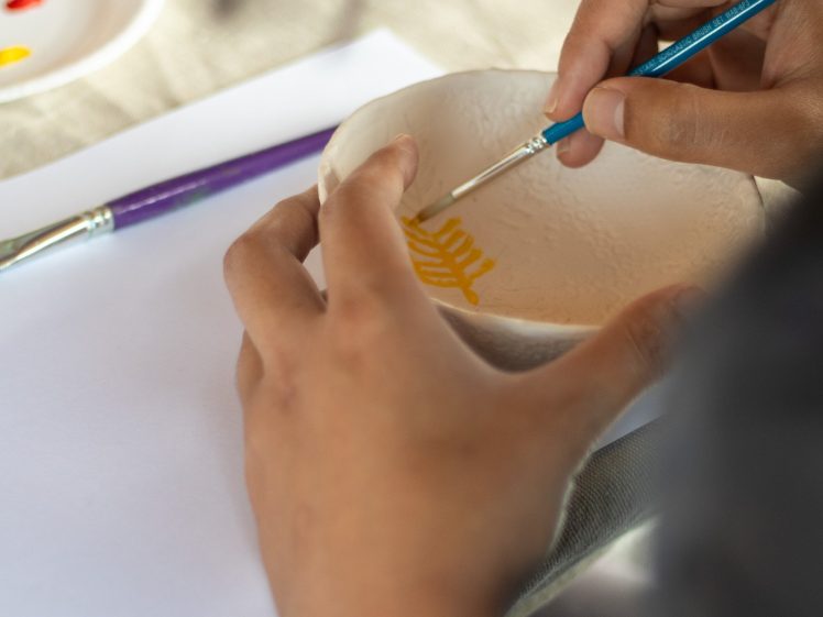 Image of A person carefully paints a yellow design on a white ceramic bowl with a thin brush. Nearby are a purple paintbrush, a piece of paper with paint, and a white sheet of paper on a beige surface.