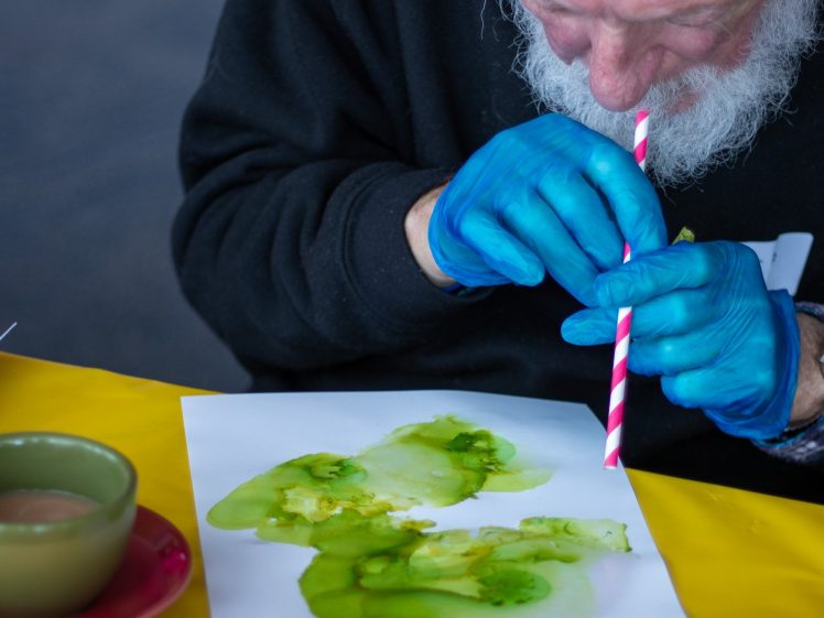 Image of An elderly person wearing blue gloves uses a red-and-white striped straw to blow green watercolour paint on a sheet of paper; a cup and saucer sit nearby on a yellow table.