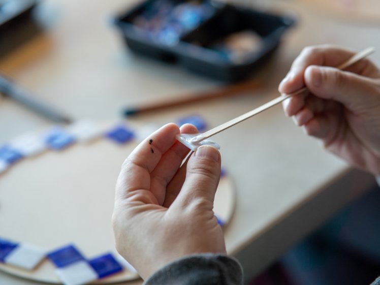 Image of A person holds a small glass tile with tweezers, preparing to place it onto a circular mosaic. Nearby are other blue and white tiles and art supplies on a table.