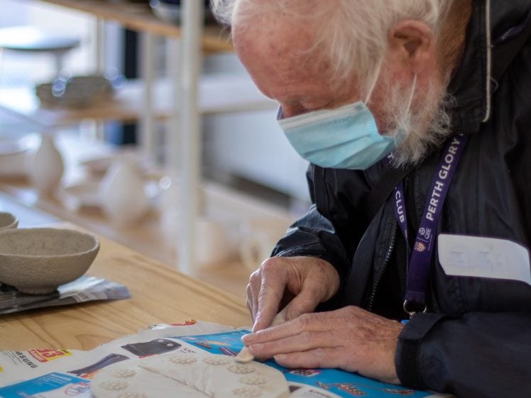Image of An elderly man with white hair, wearing a face mask and dark jacket, shapes clay on a table covered with colourful paper. Ceramic bowls and pottery are visible in the background.