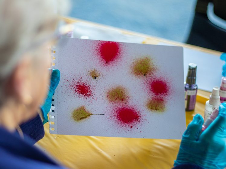 Image of A person wearing blue gloves holds a sheet of paper with colourful spray paint spots, mainly red and yellow, on a yellow table with spray bottles nearby. The focus is on the artwork.
