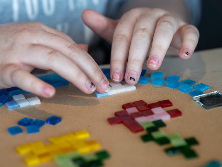 Image of Close-up of a person's hands arranging colourful mosaic tiles on a round board. The person is wearing a grey sweatshirt, and some of their fingernails are painted.