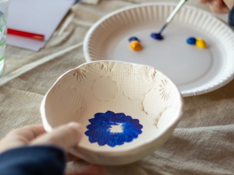Image of A person paints a white ceramic bowl with blue paint using a brush. Another hand holds the bowl. On the table are a paper plate with blue and yellow paint, a paintbrush, and a cup of water.