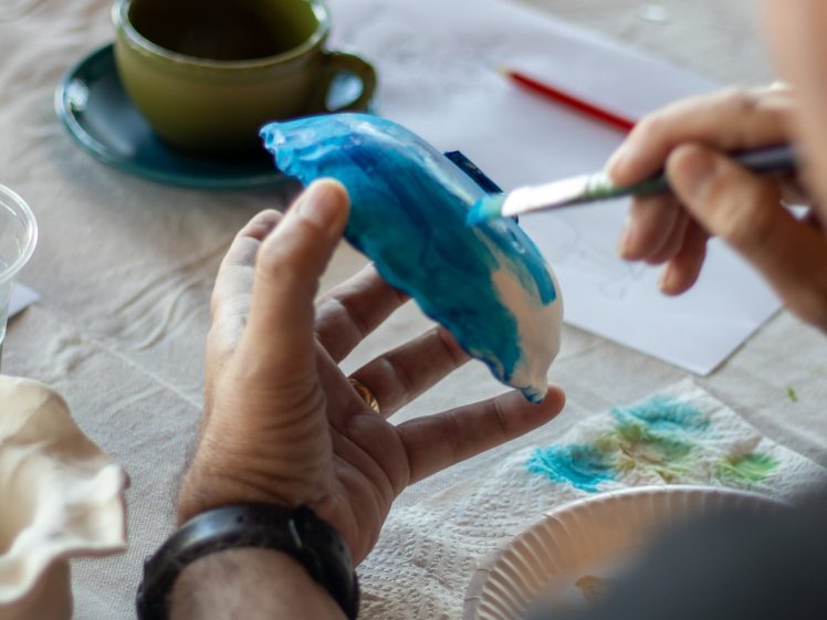 Image of A person paints a ceramic object blue whilst holding it in one hand. A paintbrush, paint, a green cup and saucer, and papers are on the table in the background.