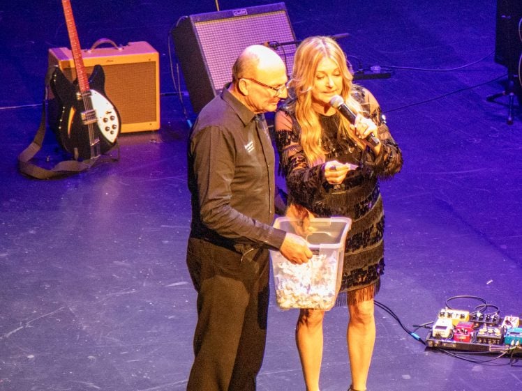 Image of A man holds a clear box of paper slips while a woman in a black dress speaks into a microphone on stage. Musical equipment, including a guitar and amplifier, is visible in the background.