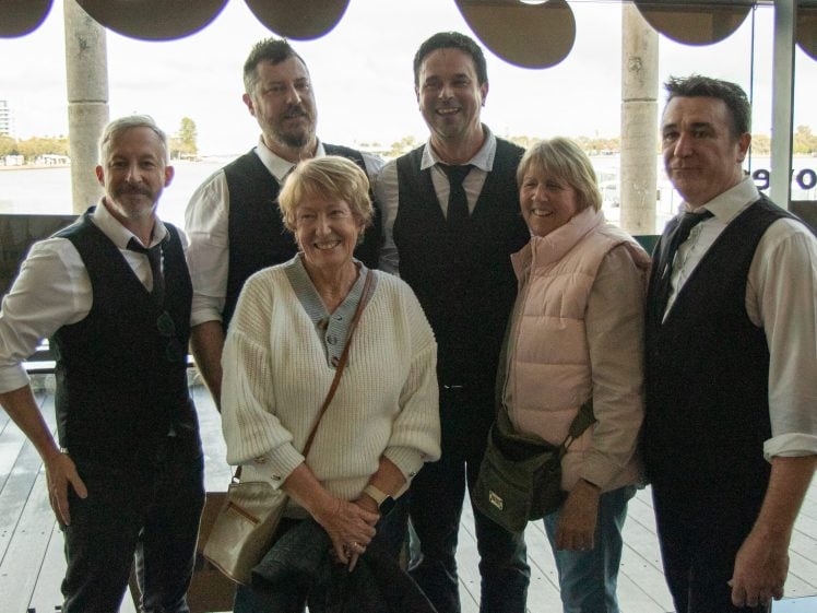 Image of Six adults, three men in waistcoats and ties standing behind two women in casual clothes, pose together indoors by large windows with a scenic riverside view in the background.