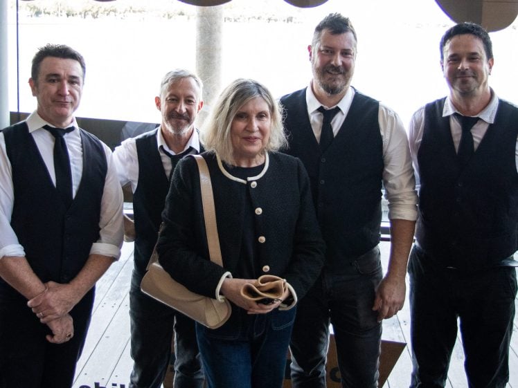Image of Five adults posing for a photo; four men in white shirts, black waistcoats, and ties stand behind a woman with shoulder-length hair holding a tan bag in front of her. They are on a wooden decking with water in the background.