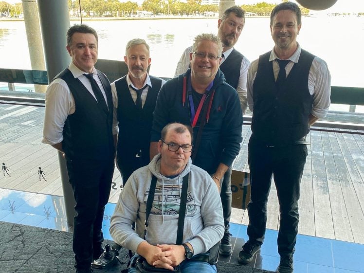 Image of Six men pose together indoors near a large window overlooking water; five are standing, dressed in black waistcoats and ties, while one man in a hoodie sits in a wheelchair in front.
