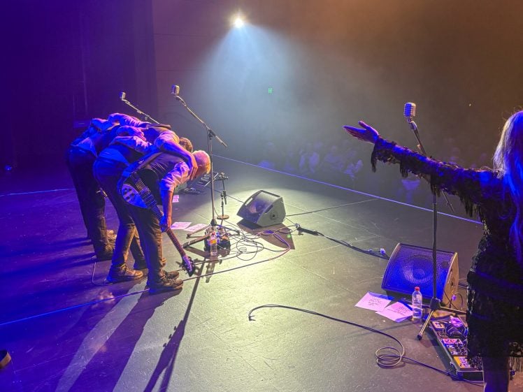 Image of Four musicians take a bow on stage under bright lights, facing an applauding audience. A performer at the right extends an arm towards the crowd; microphones and equipment are visible on the floor.