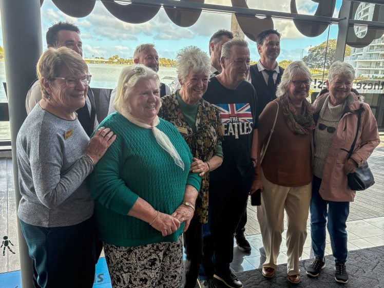 Image of A group of older adults, including several women and a man in a Beatles T-shirt, smile and pose together indoors by large windows overlooking water and buildings.