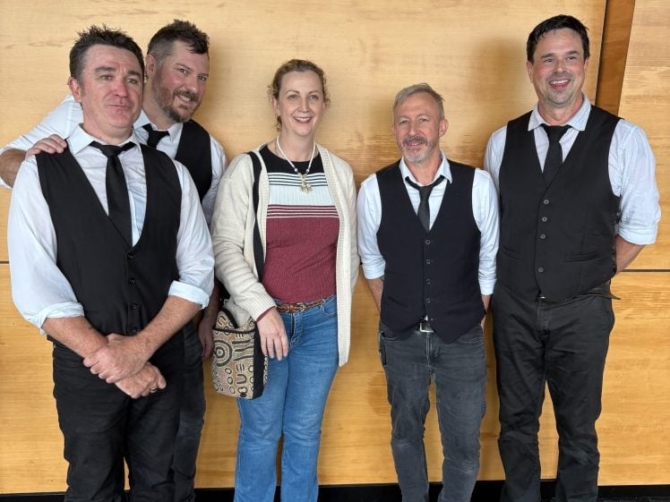 Image of Five people pose together in front of a wooden wall. Four men wear black waistcoats, white shirts, and black ties, while one woman in casual clothes stands in the centre smiling.