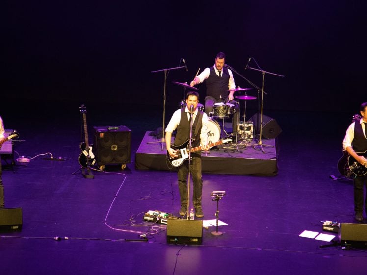 Image of Four musicians in matching outfits perform on stage with guitars, microphones, and a drum kit. They are illuminated by stage lights against a dark background. The drummer is slightly elevated behind the three guitarists.
