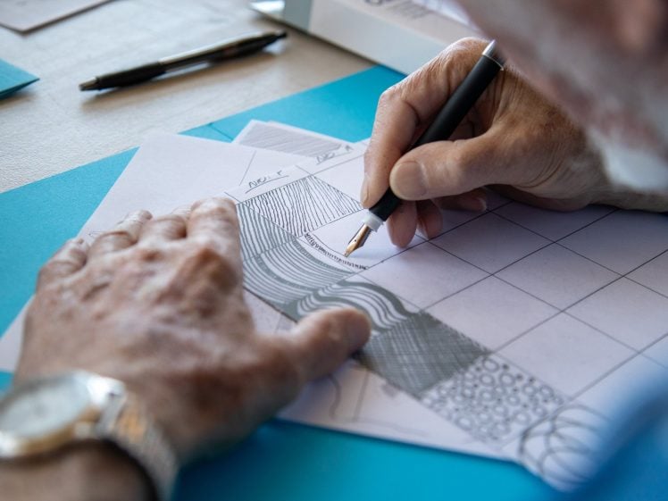 Image of A person with a watch on their wrist draws patterns with a pen on a piece of paper divided into a grid, with various intricate designs filling some of the sections. A pen and blue folder are visible on the table.