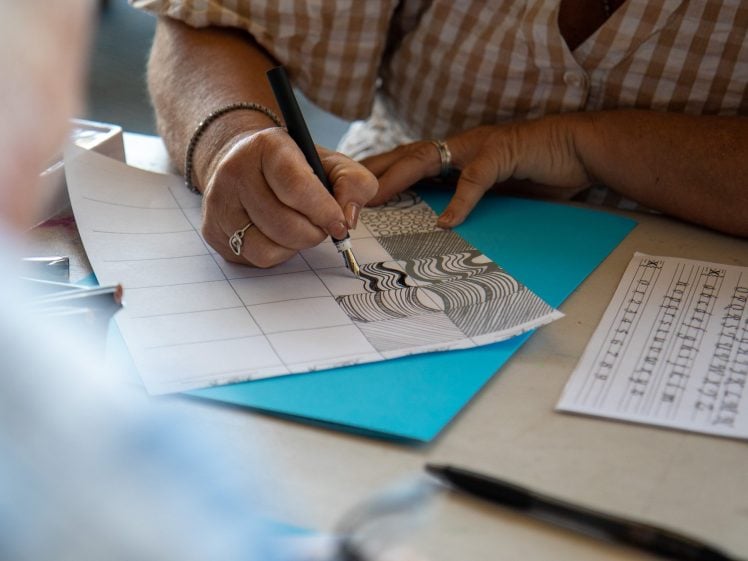 Image of A person in a checked shirt draws intricate black ink patterns on squared paper, with practice sheets, a blue folder, and pens on the table nearby.