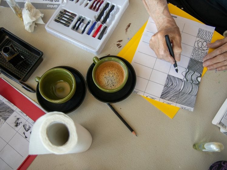 Image of A person draws patterns on a grid sheet with a pen at a table holding art supplies, two cups of coffee, kitchen roll, and drawing tools. The scene is viewed from above.
