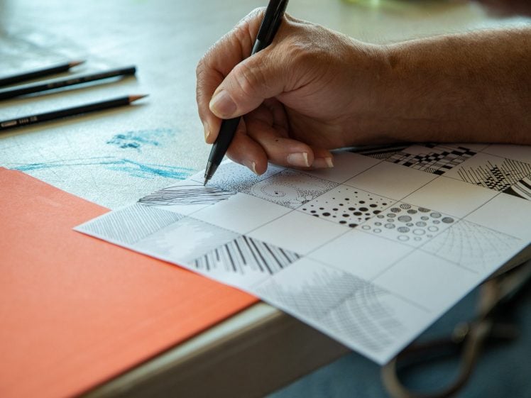 Image of A hand draws patterns with a pen on a sheet of paper divided into small squares, each featuring different designs. Three pencils and an orange folder are on the table nearby.