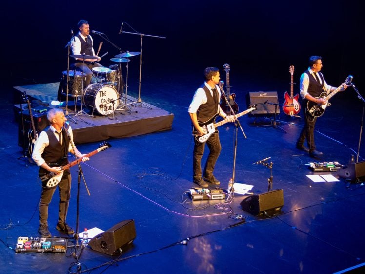 Image of Four musicians dressed in matching black waistcoats and white shirts perform on stage with guitars and drums. They stand under stage lights with microphones, amplifiers, and pedalboards visible. A drum kit labelled The Bootleg Beatles is at the back.
