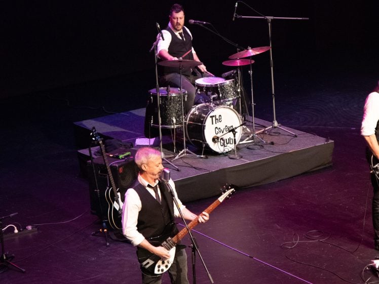 Image of A band performs on stage; a drummer plays a drum kit labelled The Cavern Club while a guitarist sings into a microphone. Both musicians wear white shirts and black waistcoats. The stage is lit with purple-tinted lights.