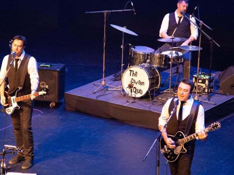 Image of Three musicians in black waistcoats and white shirts perform on stage. One plays bass and sings, another plays electric guitar and sings, and the third plays drums behind a kit labelled The Cavern Club.