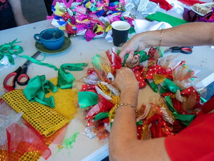 Image of A person ties colourful ribbons and mesh onto a wreath at a craft table scattered with fabric, scissors, coffee cups, and other craft supplies.
