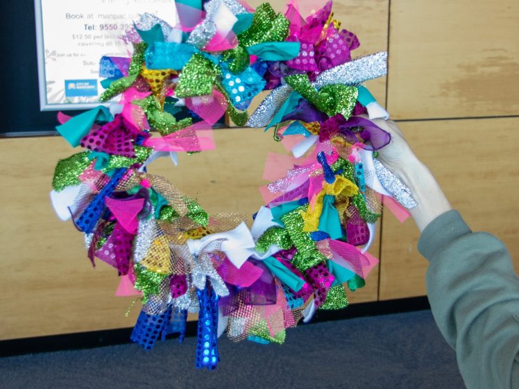 Image of A person holds a colourful wreath made of various textured and patterned fabric pieces, including glittery gold, green, pink, blue, and purple, against a background of wood-panelled walls and a posted sign.
