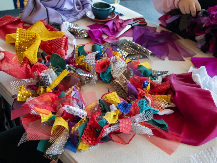 Image of Several hands work with colourful fabric scraps, tulle, and sequins on a table, creating textured, vibrant collages. Scissors, a teacup, and more fabric pieces are also visible.
