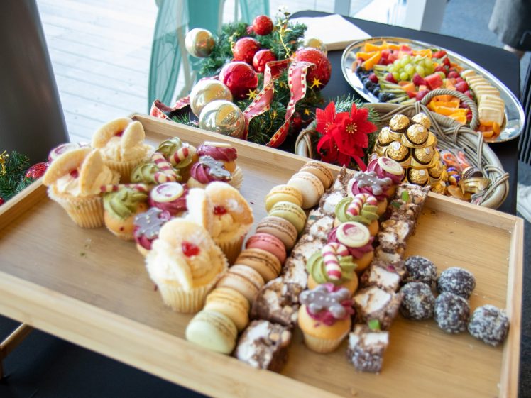 Image of A table with assorted desserts, including fairy cakes, macarons, fruit tartlets, brownies, and chocolate truffles, displayed on wooden trays. There are also fruit platters and Christmas-themed decorations in the background.