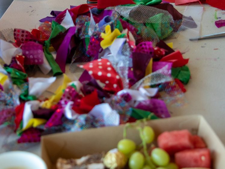 Image of A table displays colourful fabric scraps and partially made wreaths, with hands working in the background. In the foreground, there are snacks including grapes, pastries, and a cup of coffee.