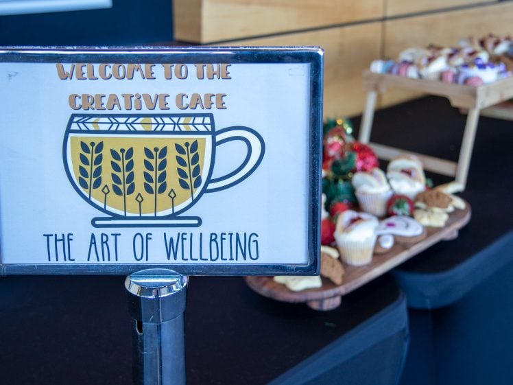 Image of A sign reading Welcome to the Creative Café: The Art of Wellbeing is displayed in front of a table with assorted pastries, cupcakes, and holiday decorations in the background.