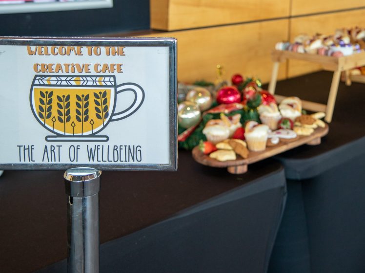 Image of A sign reading Welcome to the Creative Café: The Art of Wellbeing stands in front of a table with assorted pastries and holiday decorations.