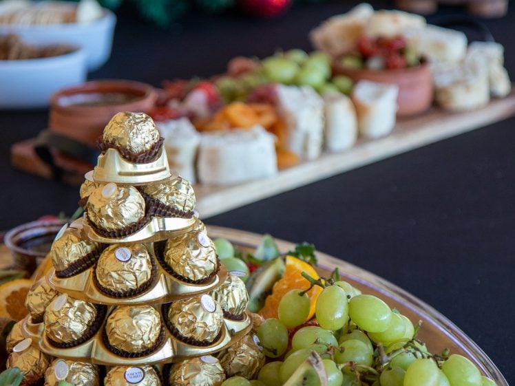 Image of A festive table display with a pyramid of Ferrero Rocher chocolates surrounded by assorted fresh fruit, including grapes, oranges, and kiwi, with more holiday treats and decorations in the blurred background.