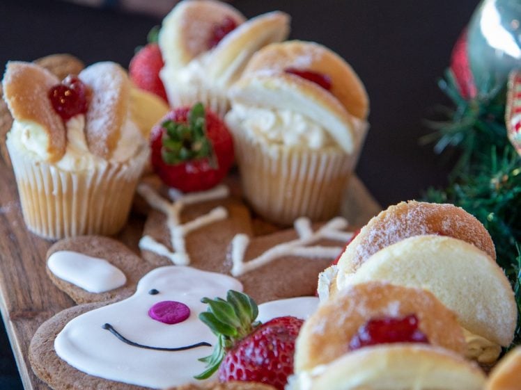Image of A wooden board with assorted pastries, cupcakes, strawberries, and a gingerbread biscuit decorated as a reindeer with white icing and a pink nose. Another platter with more food is blurred in the background.