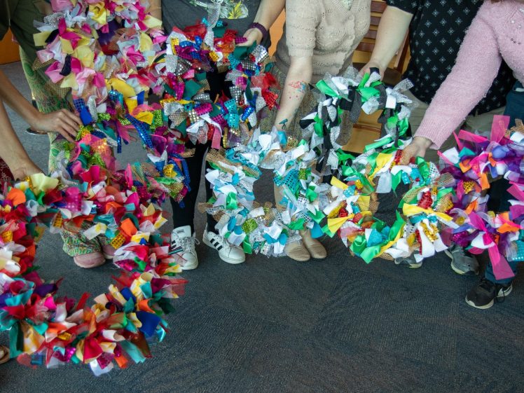 Image of Several people stand in a circle, each holding a colourful wreath made from knotted strips of fabric, while their heads are out of the frame. They are standing on a blue carpeted floor.