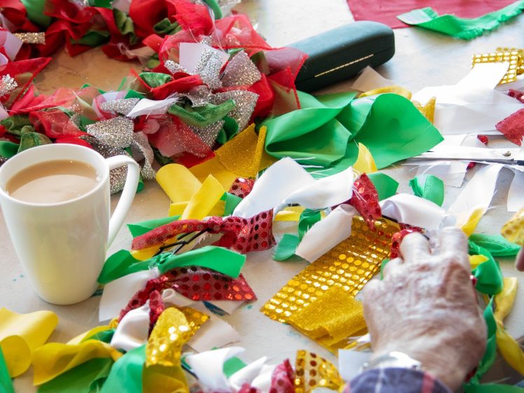 Image of Two people sit at a table making colourful wreaths from strips of red, green, yellow, and shiny fabric. Scissors and a mug of tea are on the table alongside their craft materials.